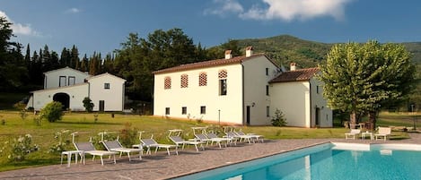 Un área de piscina con sillones reclinables, un edificio grande con techo de tejas y un paisaje montañoso de fondo.