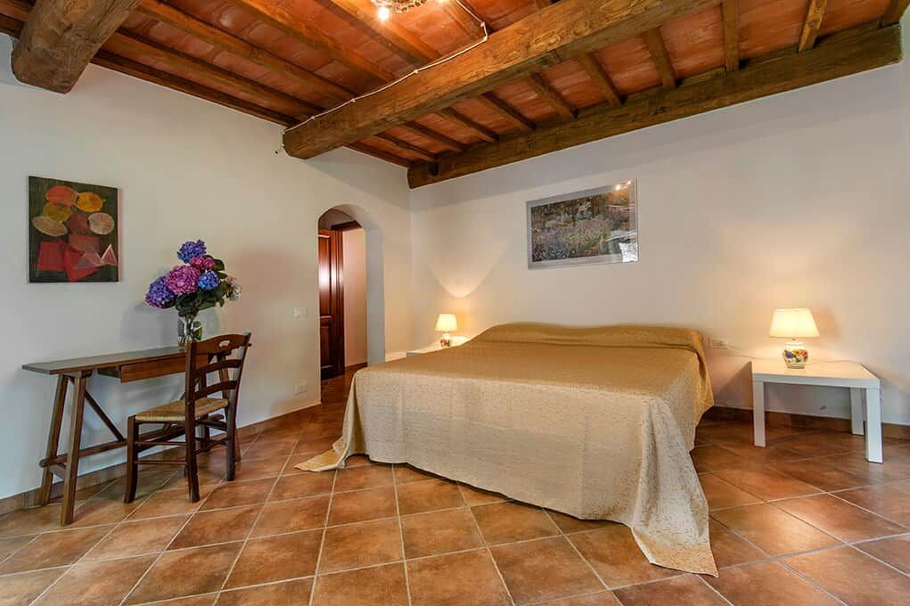 A bedroom with a bed, a desk, and chairs, a wooden ceiling, and a tiled floor.