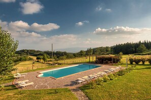 Relax by the serene pool surrounded by lush greenery and a clear sky, with lounge chairs and umbrellas for ultimate relaxation.