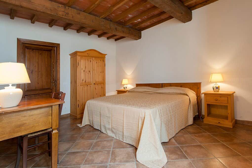 A bedroom with a bed, wooden headboard, bedside tables with lamps, a wooden wardrobe, and a desk with a lamp.