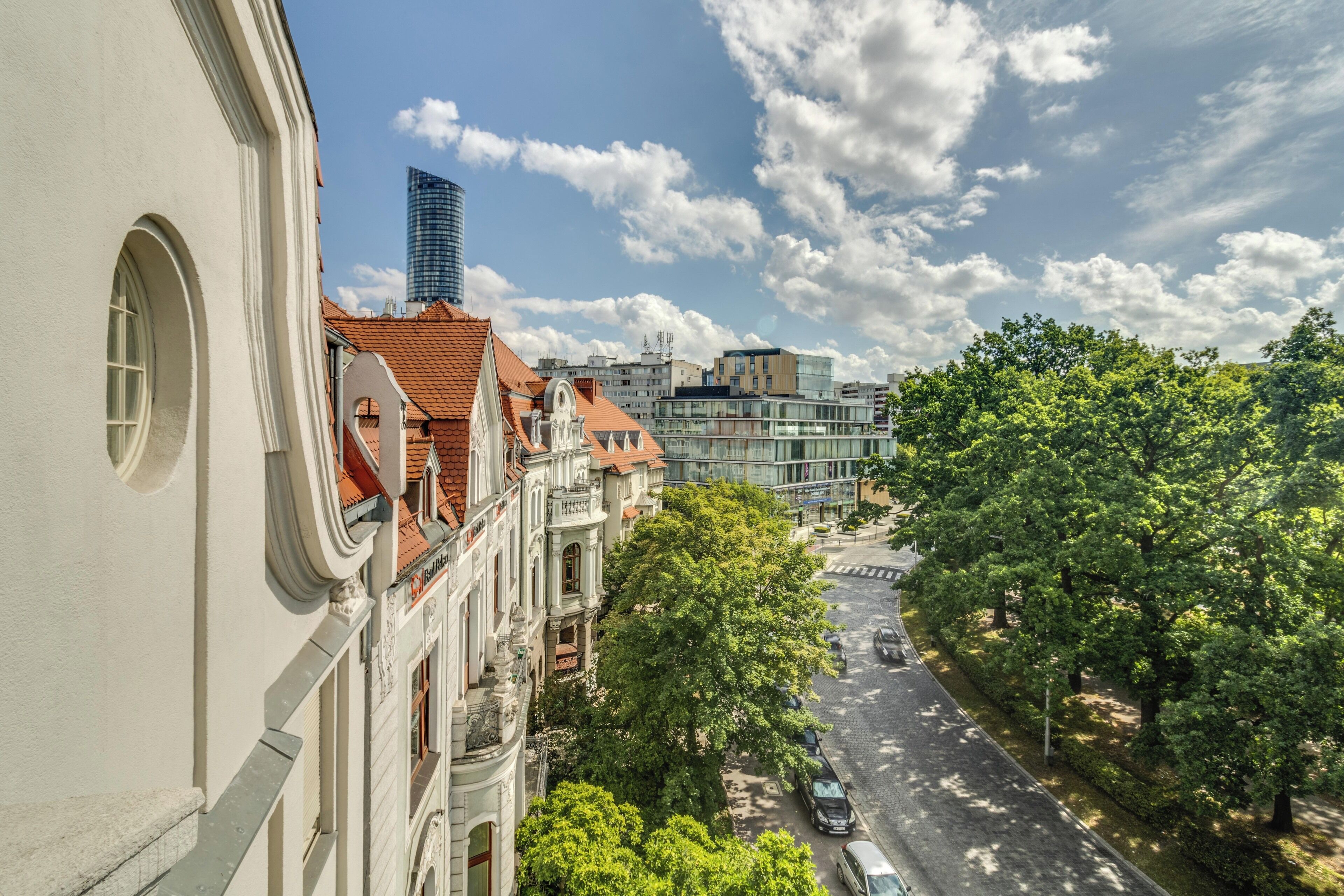 Una veduta di strada con un mix di edifici tradizionali e moderni, cielo sereno e viale alberato.