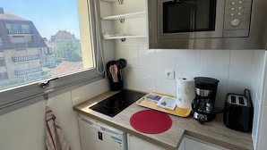 Une cuisine compacte équipée d’un four à micro-ondes, d’une machine à café et d’un grille-pain. Une fenêtre offre une vue sur les bâtiments environnants.