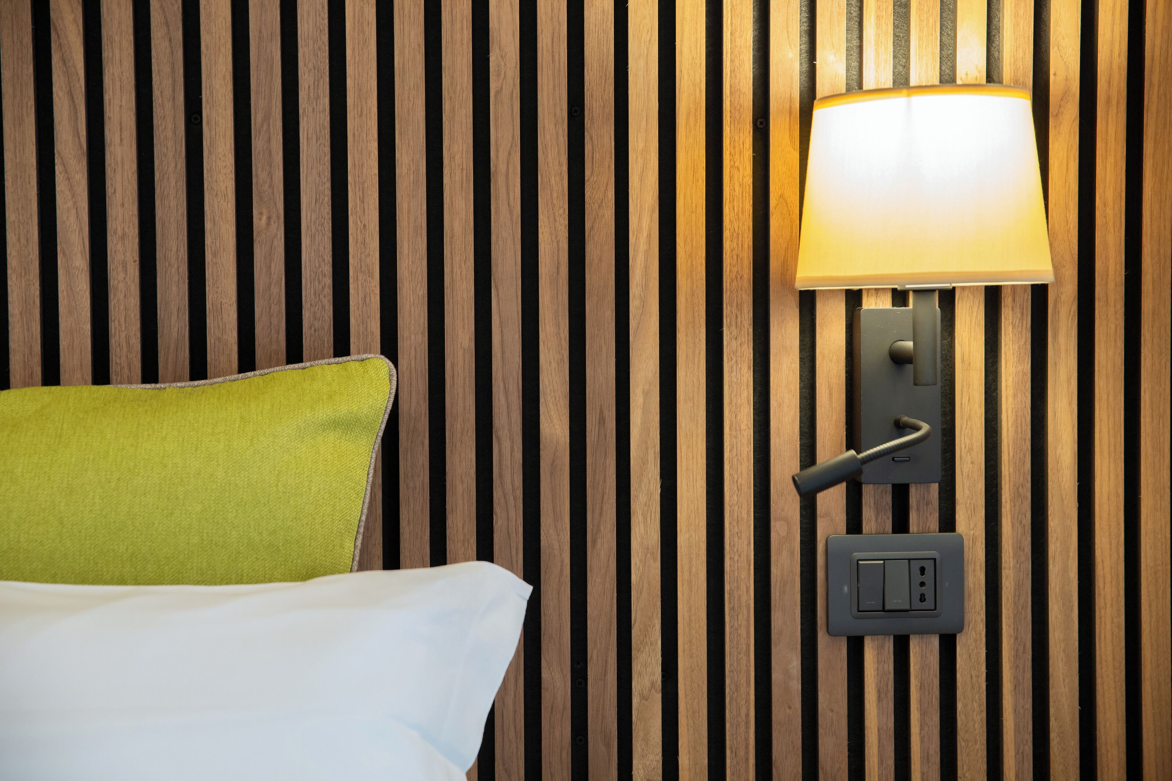 Una cama con un almohadón verde frente a una pared de listones de madera, una lámpara de pared y un enchufe.