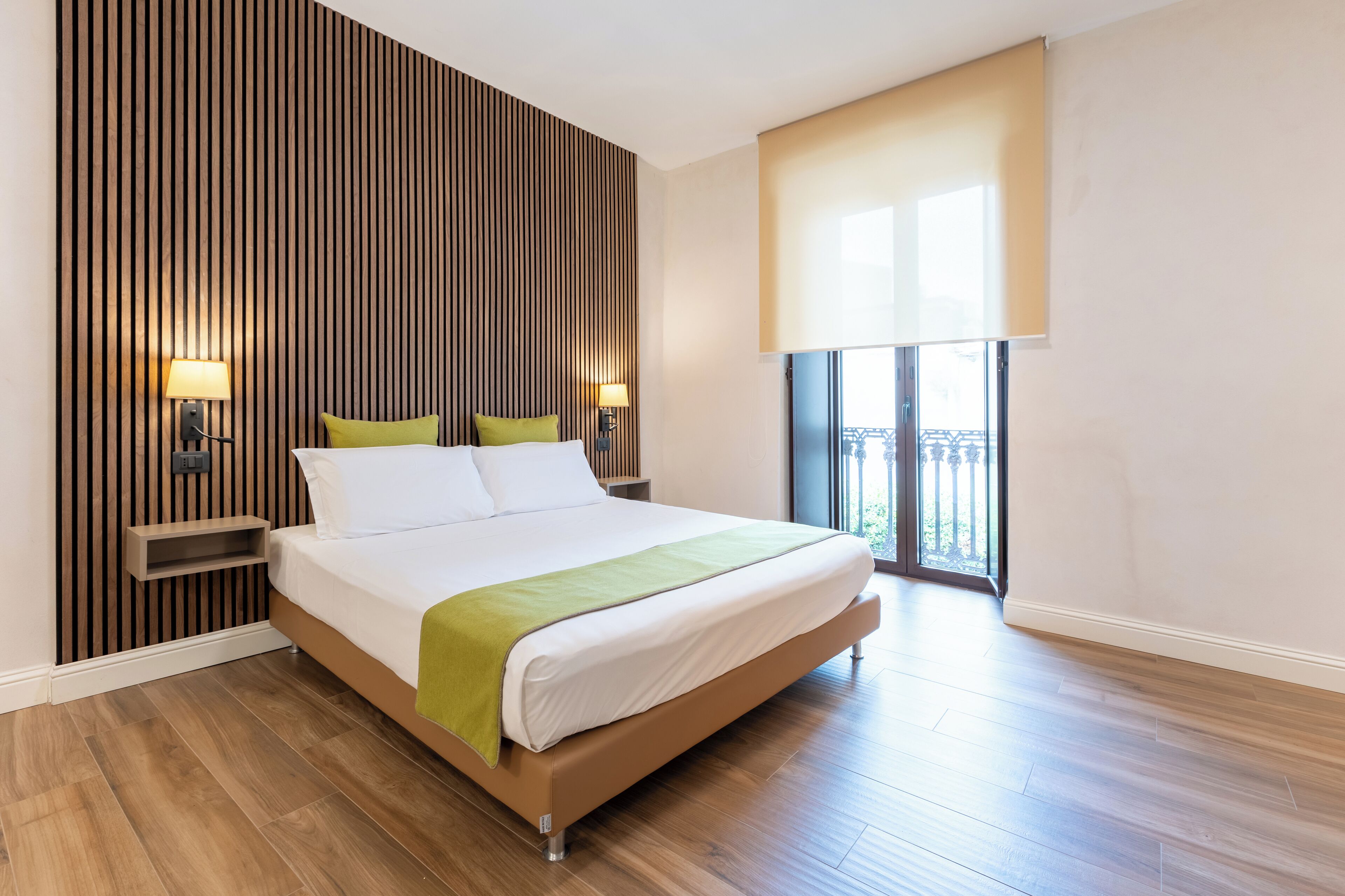 Una habitación de hotel moderna con una cama grande, un panel de pared de madera, dos lámparas de noche, una manta verde y un balcón con vistas.