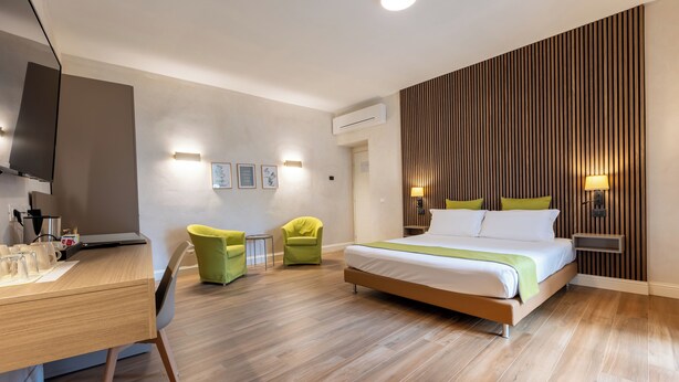 Habitación de hotel moderna con suelo de madera, cama con ropa de cama verde, escritorio con computadora portátil, sillón verde y televisor montado en la pared.