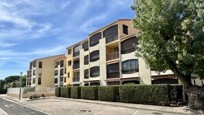 Un edificio residencial de varios pisos con balcones, un estacionamiento y un seto a lo largo de la acera.