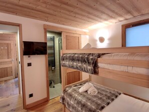 Habitación con literas, un televisor en la pared, techo de madera y una puerta que da a otra habitación.