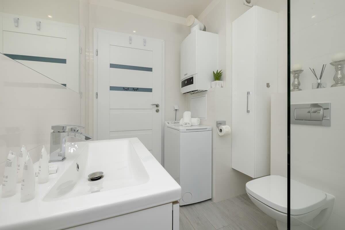Une salle de bain moderne avec un lavabo, des toilettes et une douche, le tout de couleur blanche. On trouve des articles de toilette sur le lavabo et un porte-serviettes au-dessus des toilettes.