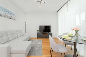 A modern living room with a grey sofa, a dining table with chairs, a TV on a black stand, and a painting on the wall.