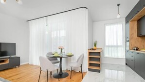 Ein modernes Wohnzimmer mit Holzboden, einem runden Esstisch mit Stühlen, einem Flachbildfernseher und einer Küchenzeile mit grauen Schränken und einer Holzablage.