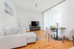 A modern living room with a sofa, a dining table with chairs, a TV on a stand, and a large window with sheer curtains.