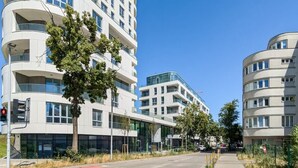 Ein modernes, mehrstöckiges Gebäude mit Balkonen und großen Fenstern, umgeben von Bäumen und blauem Himmel.