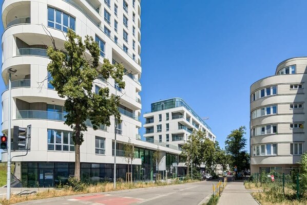 A modern multi-story building with balconies and large windows, surrounded by trees and a clear sky.