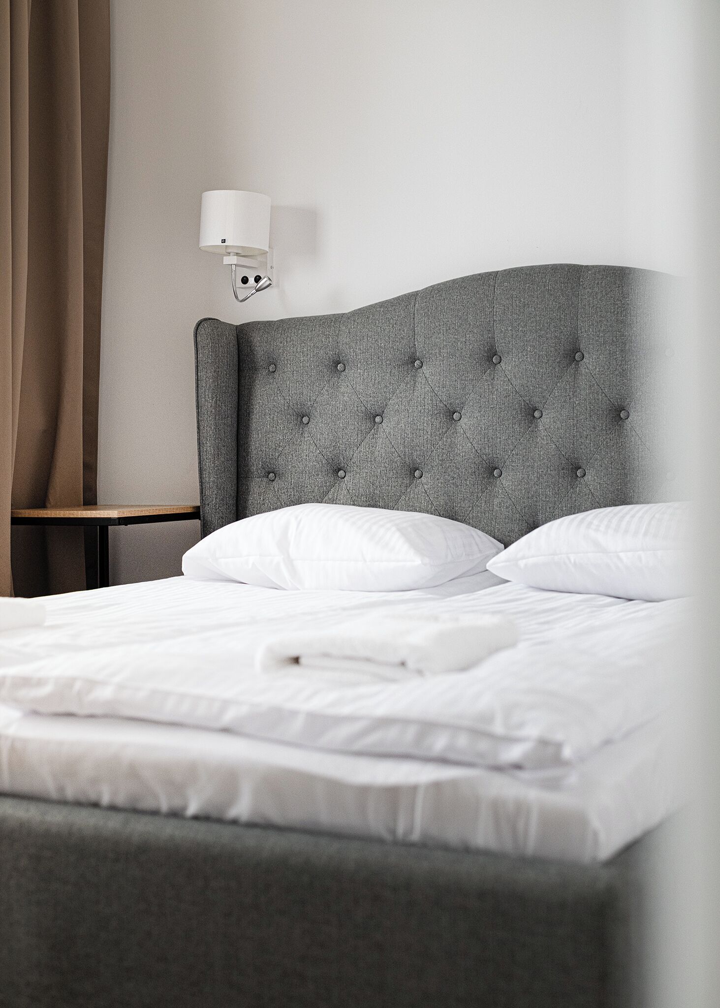 A neatly made bed with white linens, a grey tufted headboard, and a bedside table with a lamp.
