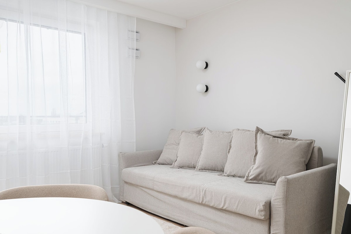 Ein schlicht eingerichtetes Wohnzimmer mit einem neutralen Sofa, einem weißen Sessel und einem großen Fenster mit leichtem Vorhang.