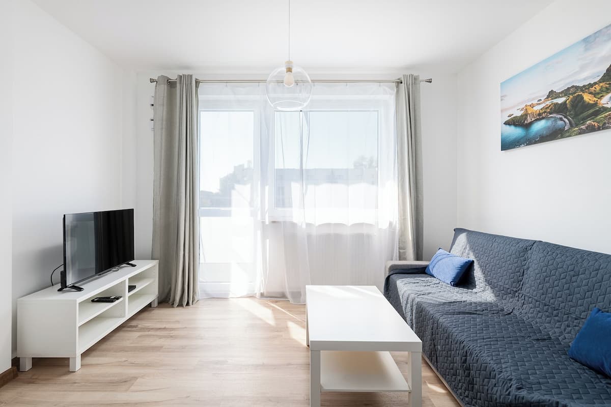 Ein modernes Wohnzimmer mit einem Flachbildfernseher auf einem weißen Standfuß, einem grauen Sofa mit blauen Kissen, einem weißen Couchtisch und einem großen Fenster mit transparenten Vorhängen.