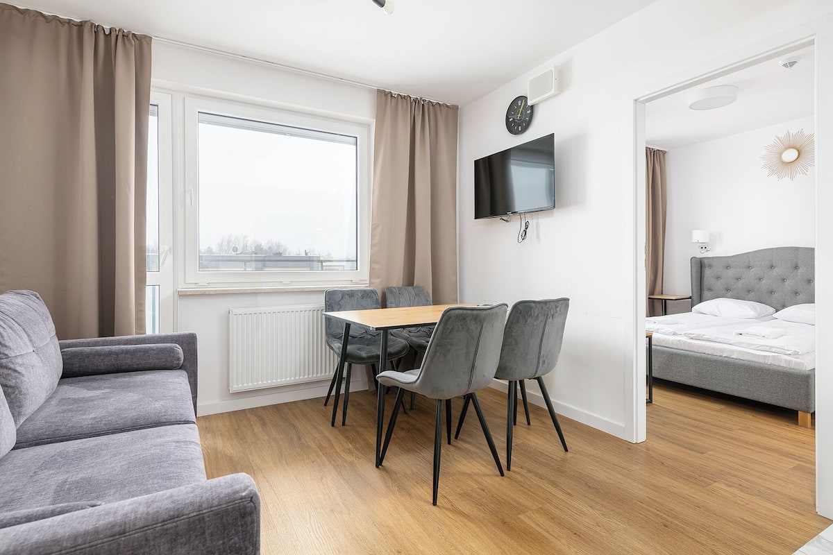 Ein modernes Zimmer mit Sofa, einem Esstisch mit Stühlen, einem an der Wand befestigten Fernseher und einem Bett mit Kopfteil.