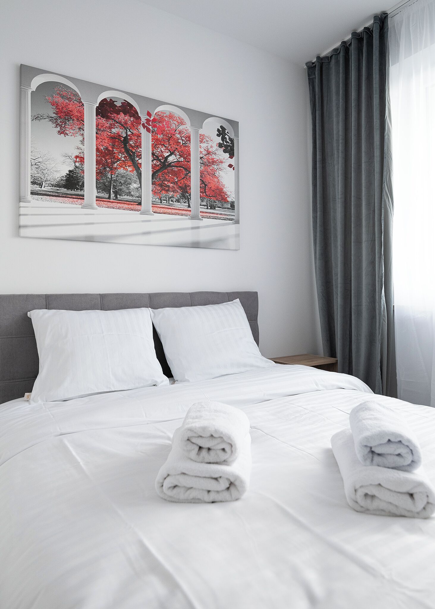 Ein ordentlich gemachtes Bett mit weißen Leinen und zwei zusammengerollten Handtüchern, ein grauer möbelbezogener Kopfteil und ein großes gerahmtes Kunstwerk mit roten Bäumen an einer weißen Wand.