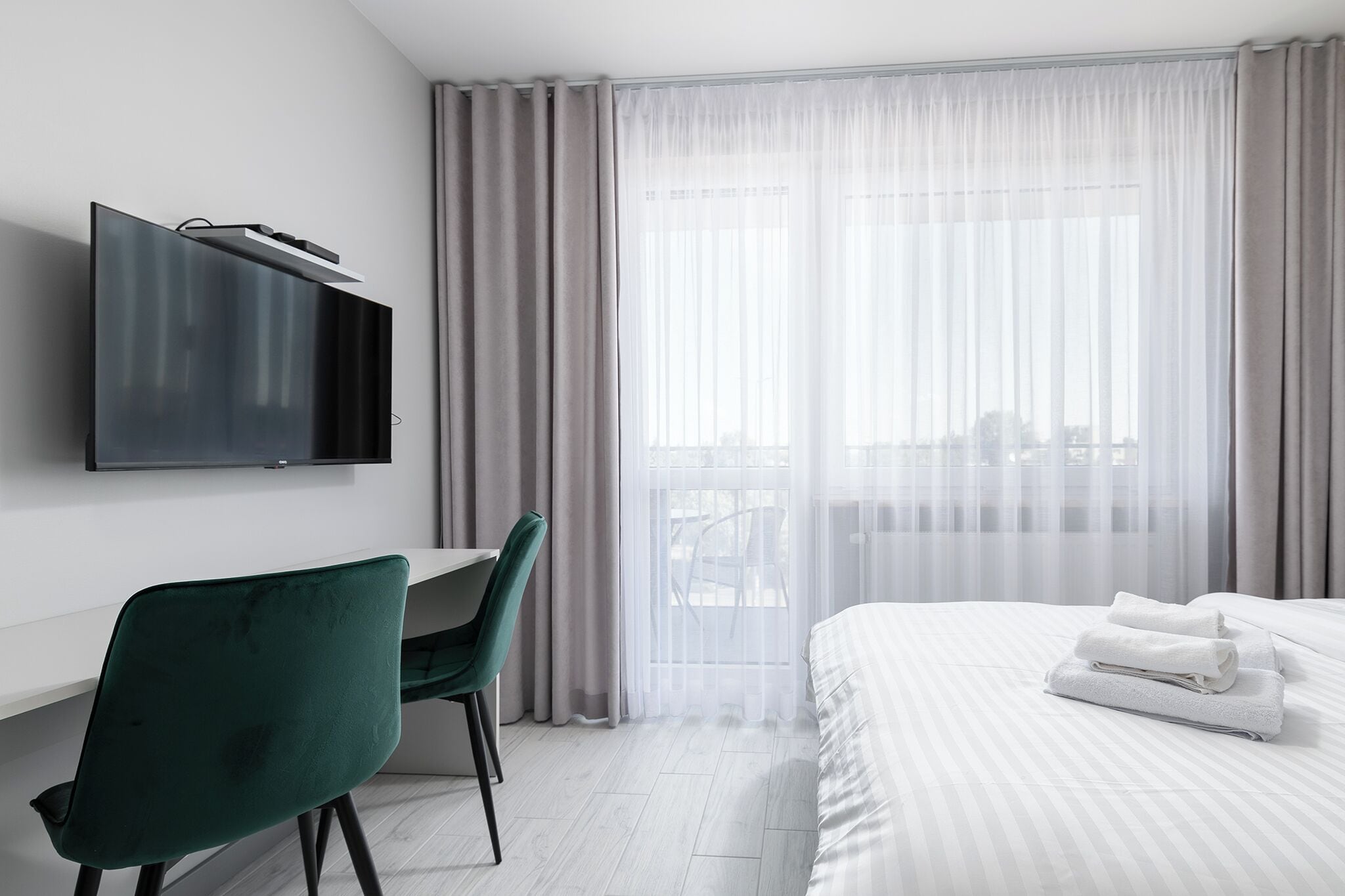 Ein modernes Hotelzimmer mit einem an der Wand befestigten Flachbildfernseher, einem Schreibtisch mit grünem Stuhl, einem ordentlich bezogenen Bett mit weißer Bettwäsche und einem großen Fenster mit durchsichtigen Vorhängen.