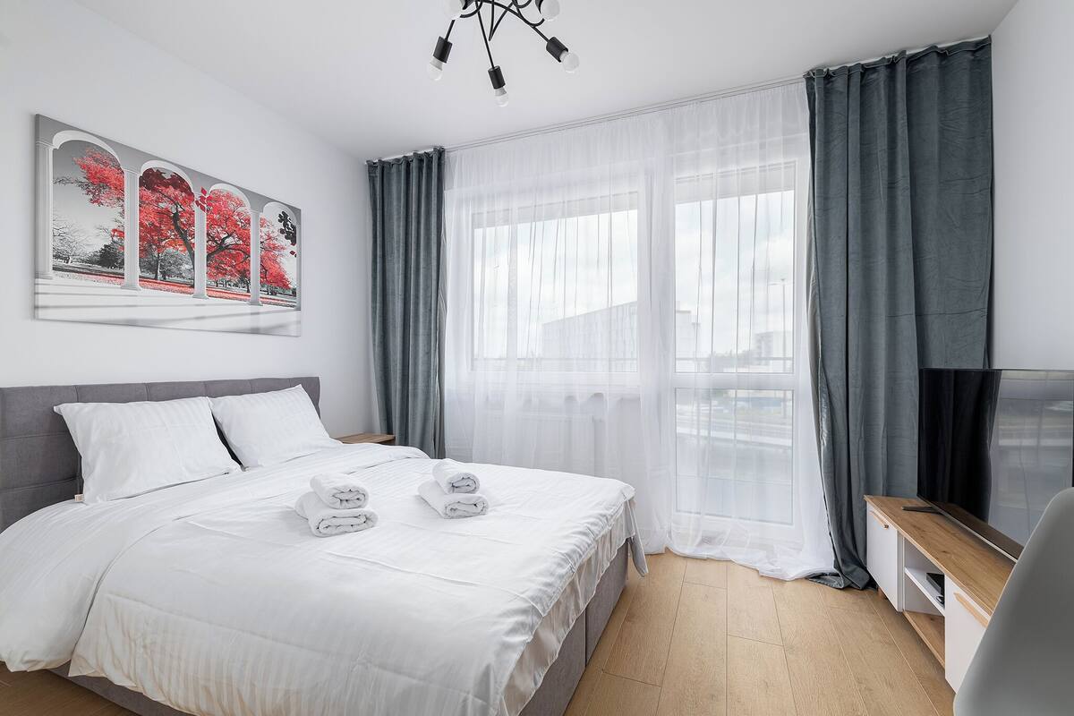 Ein modernes Hotelzimmer mit einem ordentlich bezogenen Bett, einem Flachbildfernseher und einem großen Fenster mit transparenten Vorhängen.
