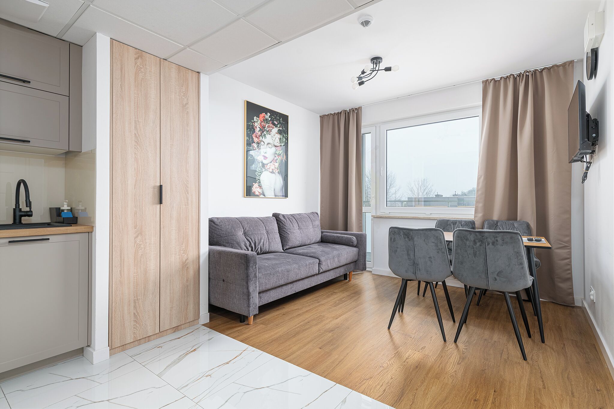 A modern apartment with a grey sofa, dining table with chairs, a kitchen area, and a large window with curtains.