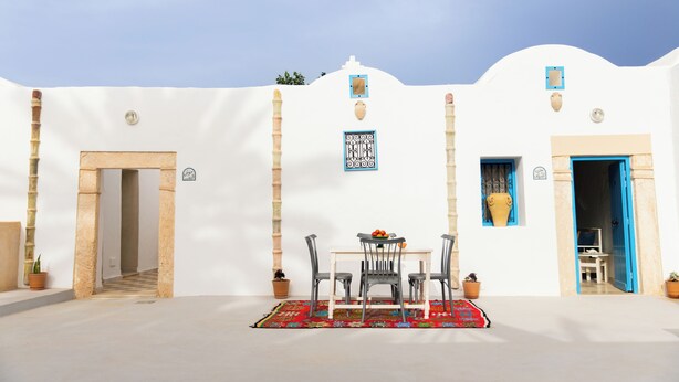 Un bâtiment blanc agrémenté de touches de bleu, avec un coin repas comprenant une table et des chaises, des plantes en pot et un tapis rouge à motifs.