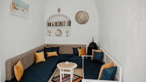 Un coin douillet avec un canapé bleu foncé, des coussins couleur moutarde, une petite table blanche et une bibliothèque remplie de livres et d’objets décoratifs.