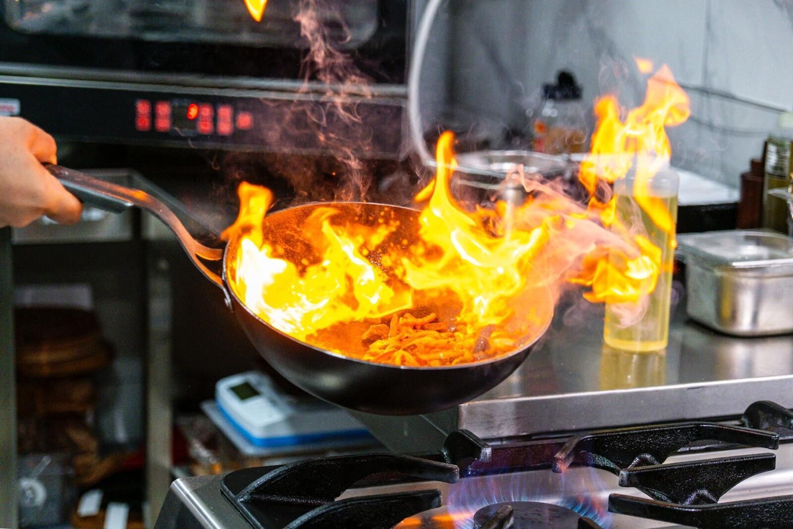 Una persona cocinando en una estufa con una sartén, con llamas visibles y utensilios de cocina al fondo.