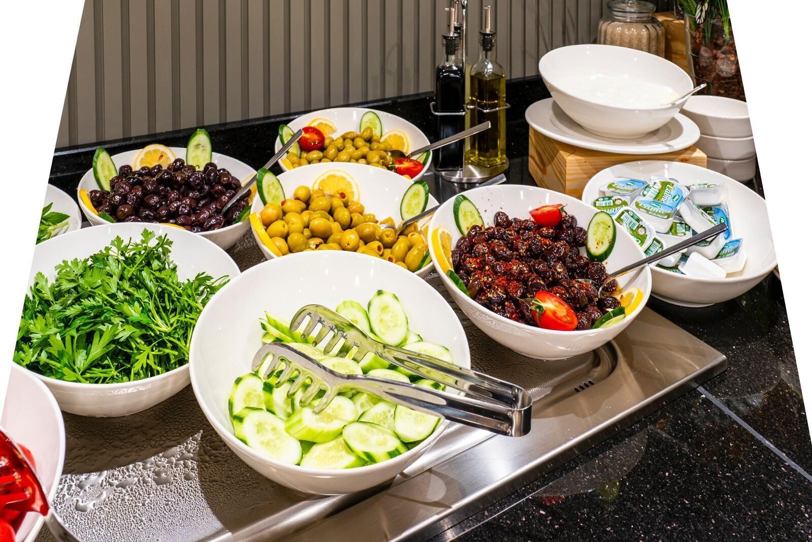 A buffet table with various dishes including olives, cucumbers, tomatoes, and a salad.
