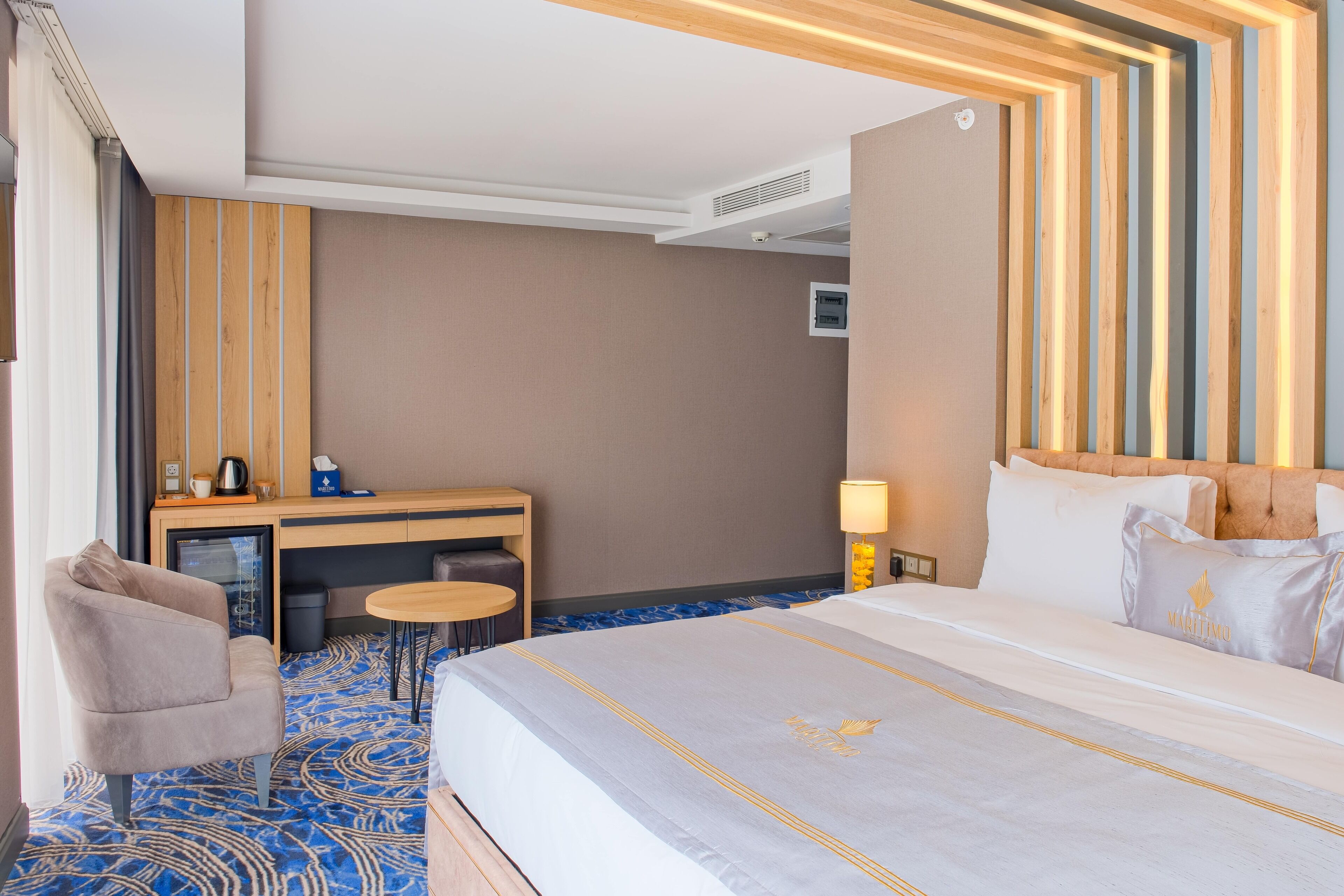A hotel room with a large bed, a desk with a chair, a small table, and a lamp.