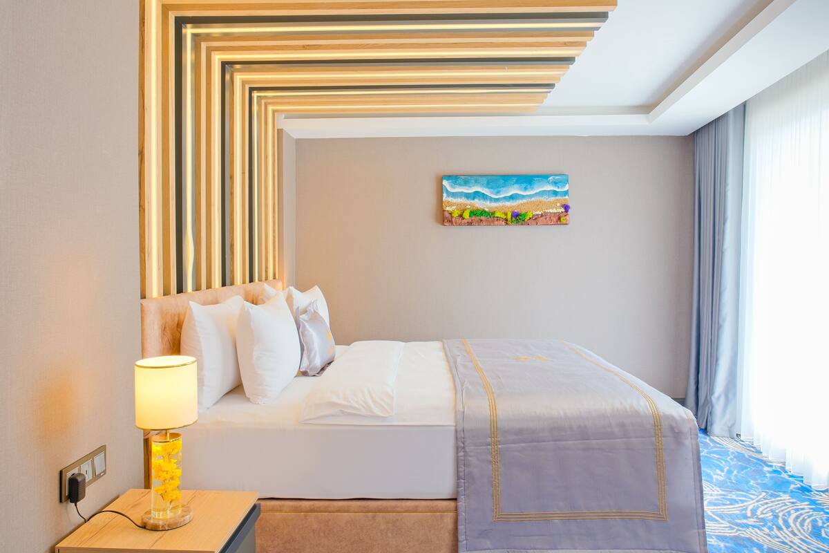 Quarto de hotel com cama, mesa de cabeceira com abajur, pintura de uma praia e janela com cortinas.