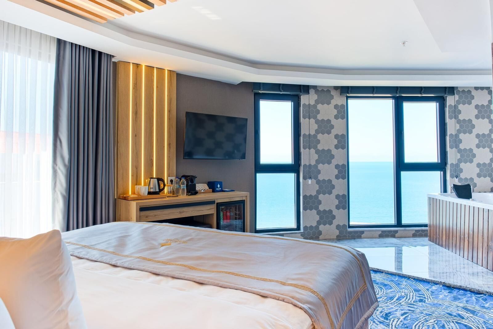 A modern hotel room with a large bed, a desk with a coffee maker, and a view of the sea through large windows.