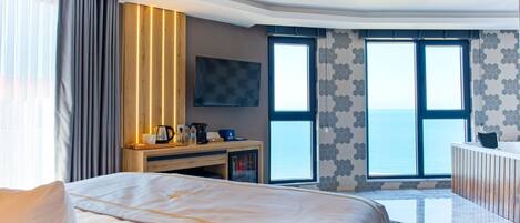A modern hotel room with a large bed, a desk with a coffee maker, and a view of the sea through large windows.