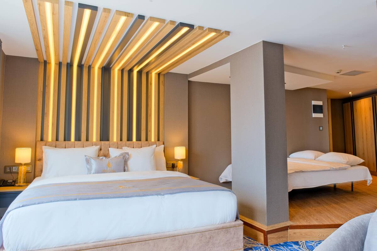 Quarto de hotel com uma cama grande, mesas de cabeceira com abajures, design de teto em madeira e uma segunda cama ao fundo.