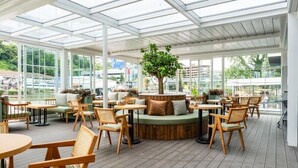 Un vaste espace intérieur aménagé avec des tables et des chaises en bois, un coin salon central et de grandes fenêtres offrant une vue sur la ville.