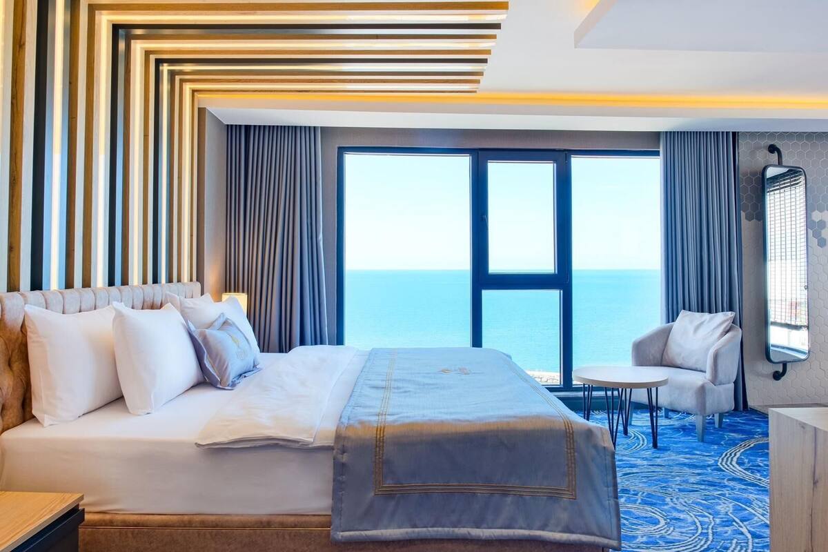 Quarto de hotel com uma cama grande, uma mesinha, uma cadeira e vista para o mar.