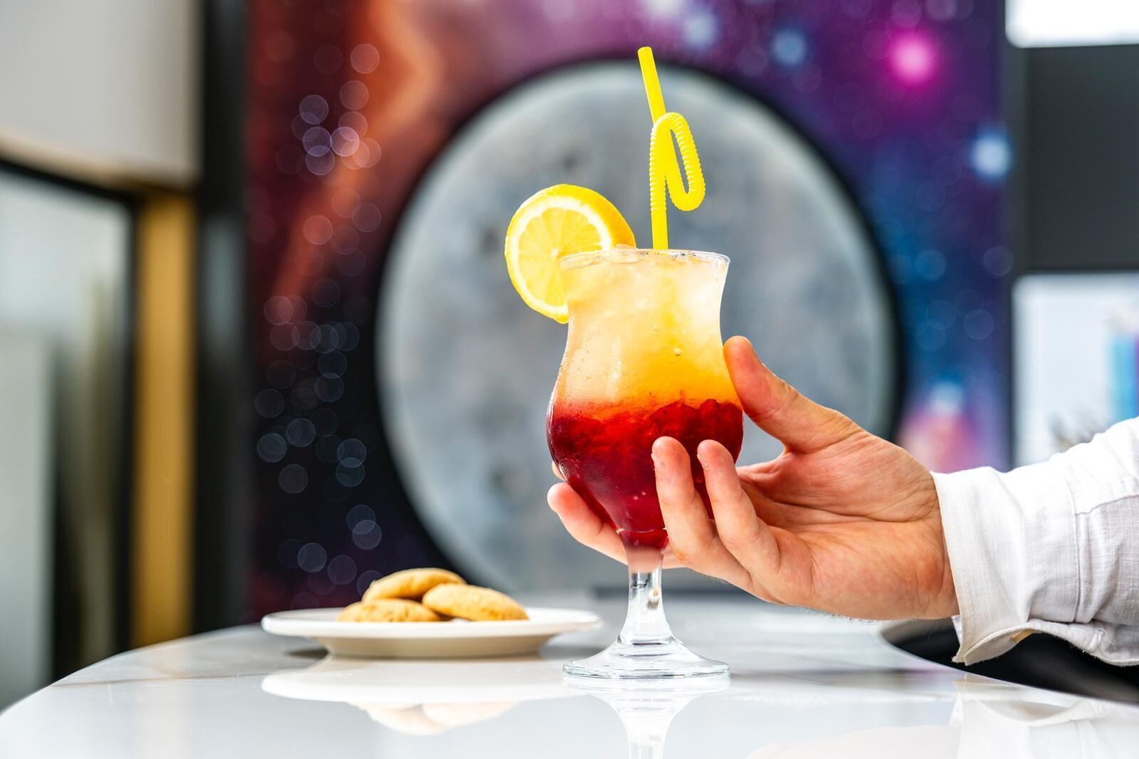 A hand holding a cocktail glass with a gradient color, a lemon slice, and a yellow straw.