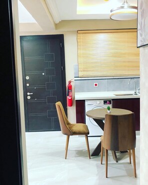 A modern kitchen with a built-in oven, a sink, and a window with blinds. There is a small round table with a chair in front of it. - UDA APARTMENT (Accra)