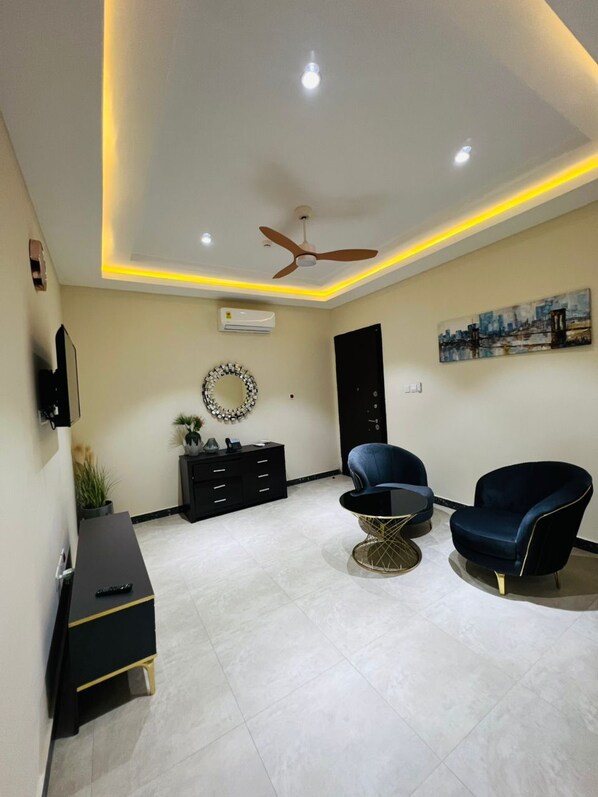 A modern living room with a ceiling fan, a black entertainment unit, and two blue chairs. - UDA APARTMENT (Accra)