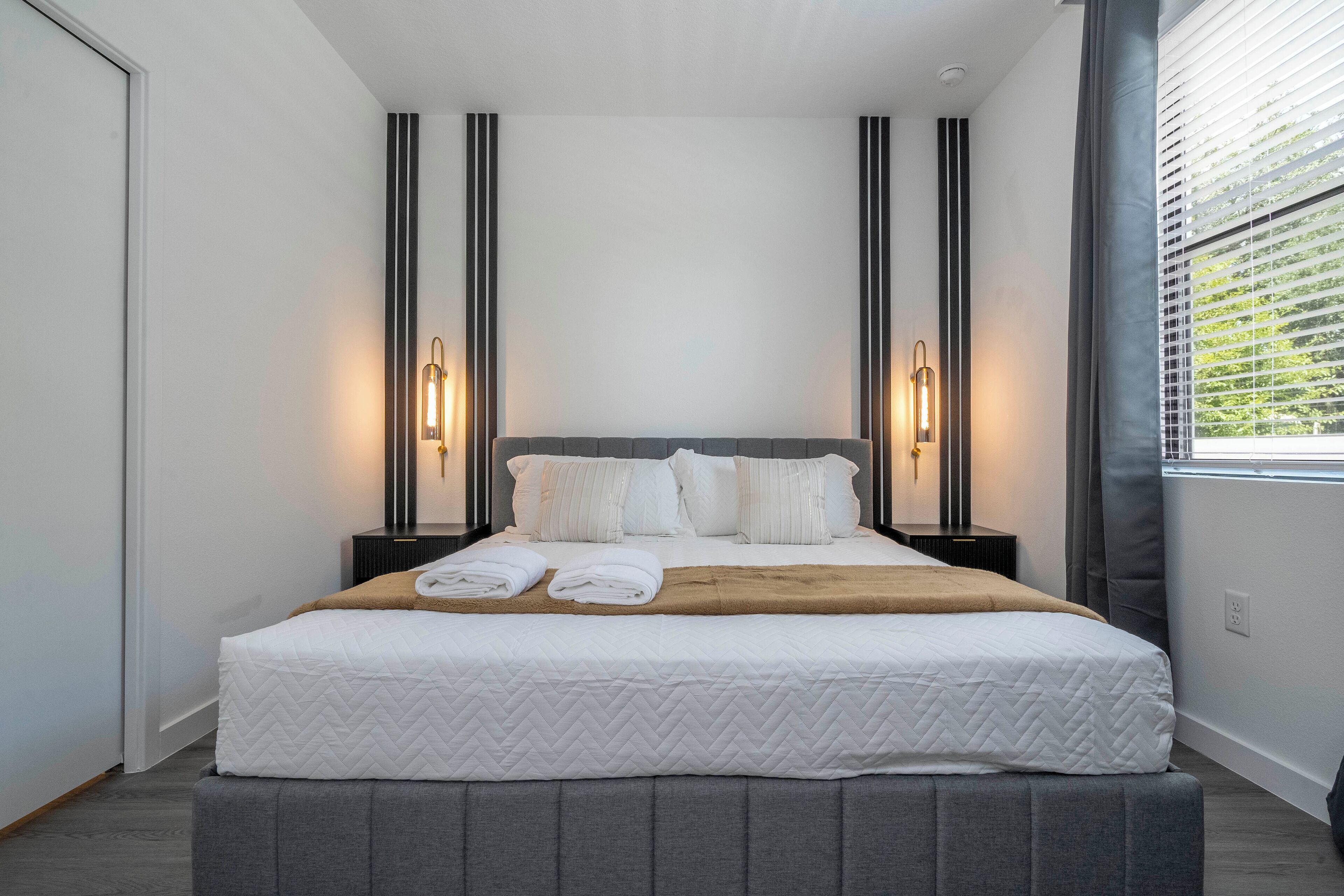 A neatly made bed with a grey headboard, two bedside tables with lamps, and a window with blinds.