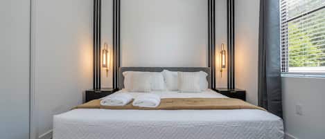 A neatly made bed with a grey headboard, two bedside tables with lamps, and a window with blinds.