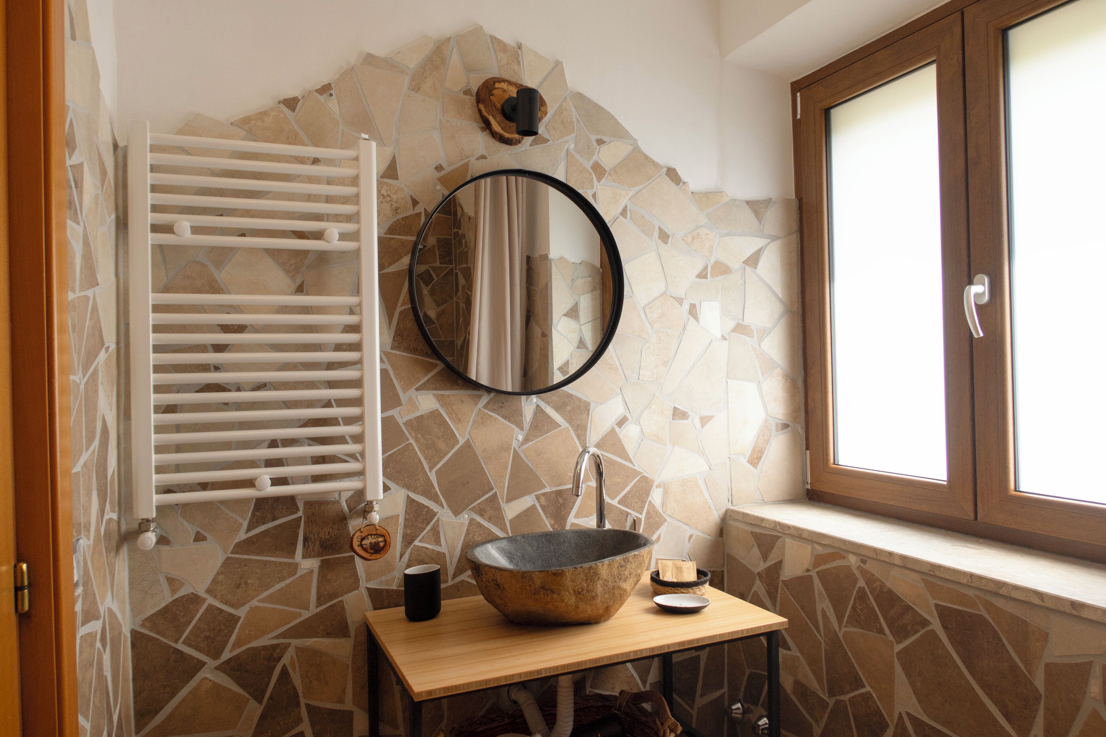 Un baño con pared de mosaico de piedra, tocador de madera, espejo redondo y ventana con contraventanas de madera.
