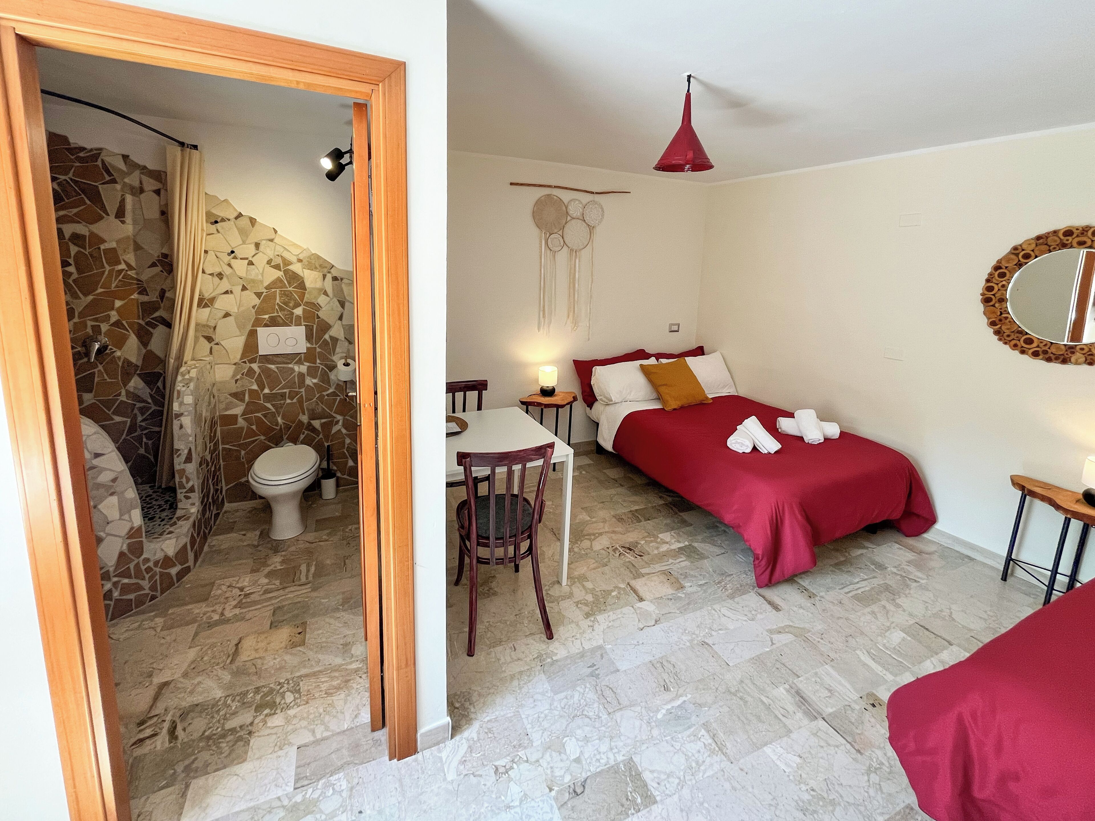 A bathroom with a mosaic wall, a shower, and a toilet. The bedroom has a bed with red bedding, a bedside table, and a mirror with a wooden frame.