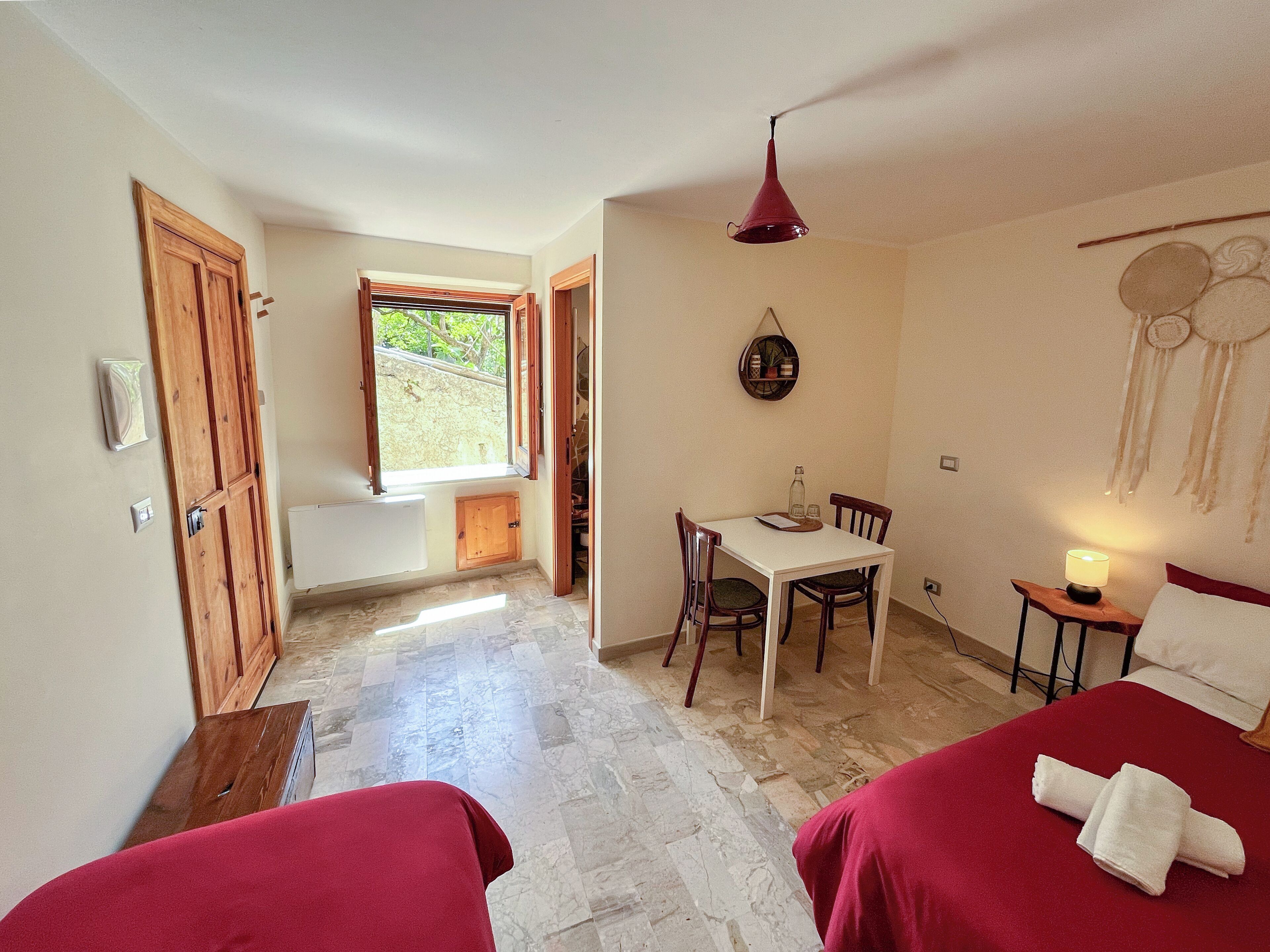 A room with two beds, a dining table with chairs, a small table with a lamp, a window with a view of greenery, and a wall with a dreamcatcher.