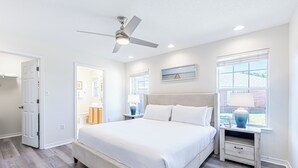 Une chambre à coucher avec un grand lit, un ventilateur de plafond, une table de chevet avec une lampe et une fenêtre avec des stores.