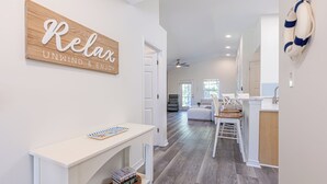 Un vaste espace de vie comprenant un meuble console blanc, un mur d’accent en bois orné d’un panneau indiquant «
Détendez-vous
», un coin repas avec une table et des chaises blanches, et un espace cuisine avec un comptoir blanc et un anneau de sauvetage accroché au mur.