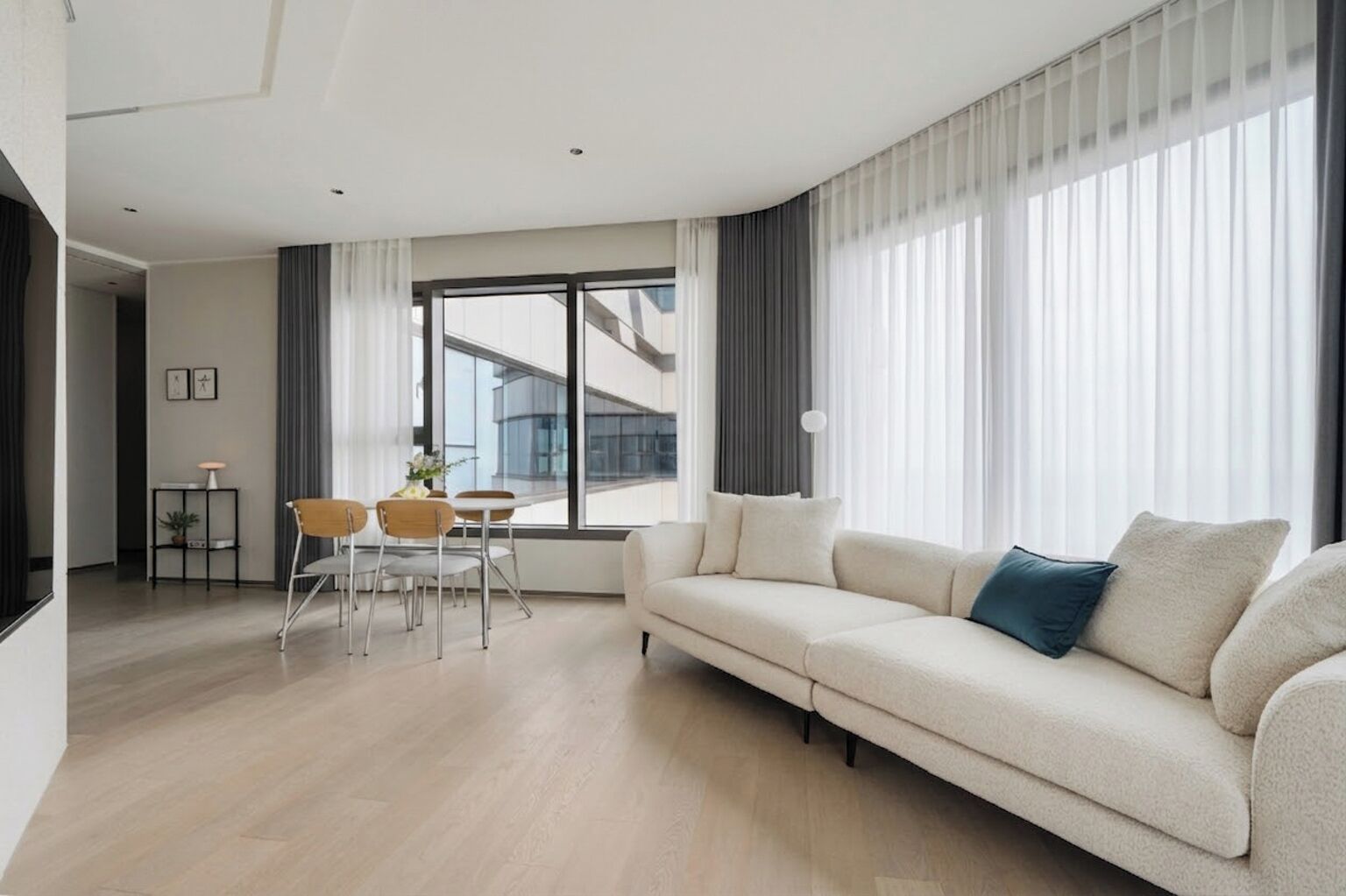 A modern living room with a sofa, a dining table with chairs, and large windows with curtains.