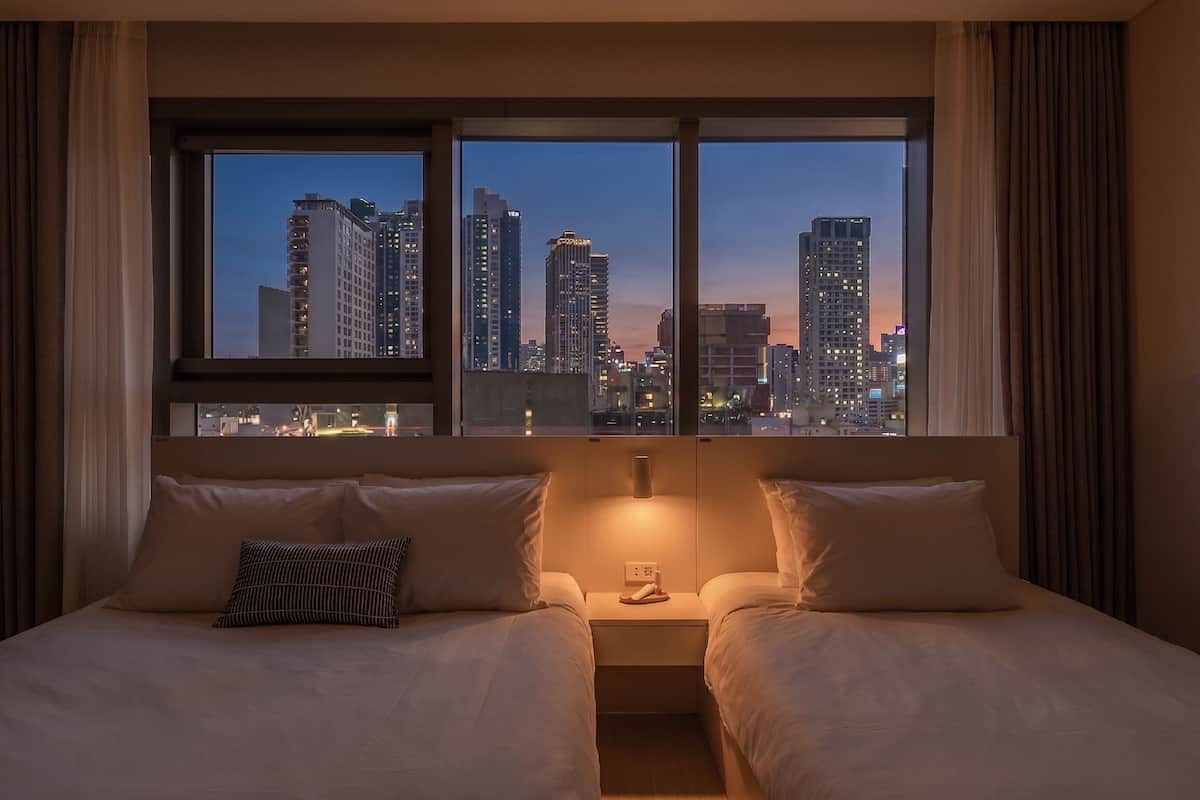 Ein Hotelzimmer mit einem Bett, zwei Kissen, einem Nachttisch, einer Lampe und einem Fenster mit Blick auf die Stadt.
