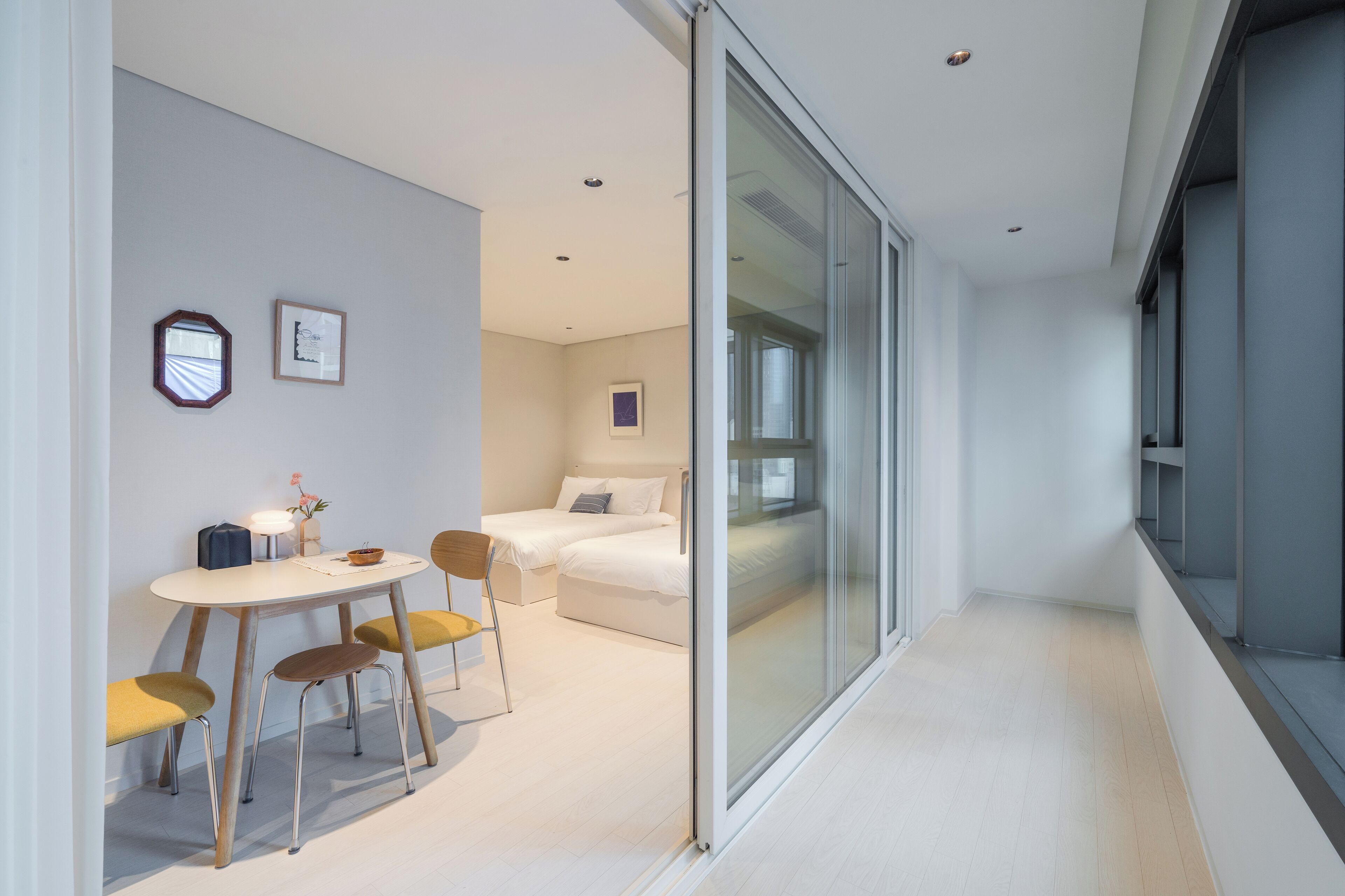 A modern hotel room with a glass partition, a bed, a small table with chairs, and a bedside table with a lamp and vase.