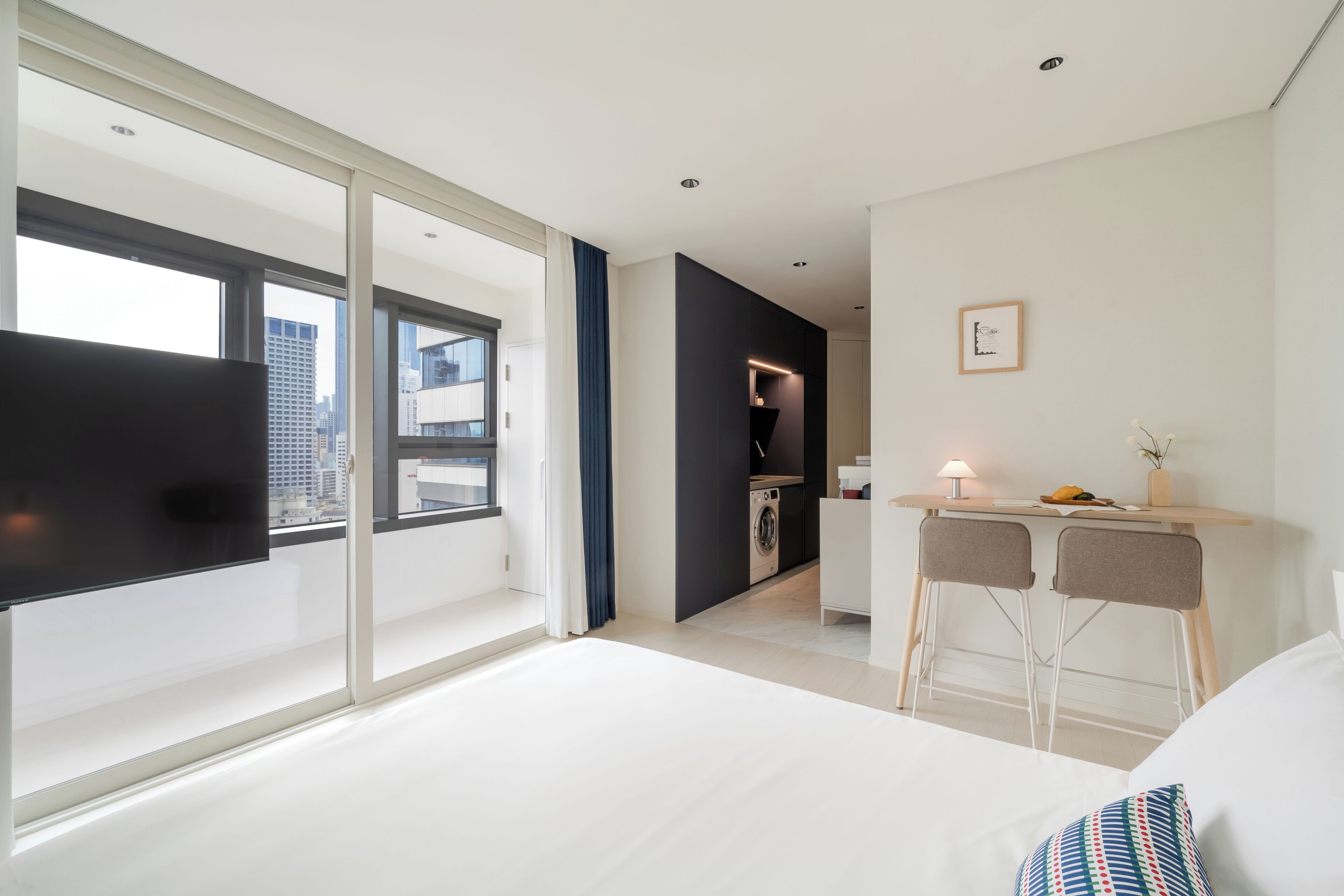 A modern hotel room with a large bed, a desk with two chairs, a small kitchen area, and a large window offering a city view.